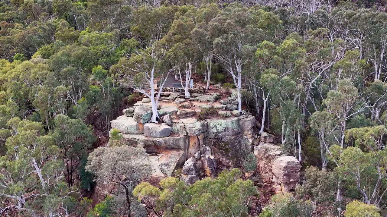 Drone camera slowly moves toward a sandstone rock formation surrounded by dense eucalyptus forest under soft, overcast daylight in Warrumbungle National Park