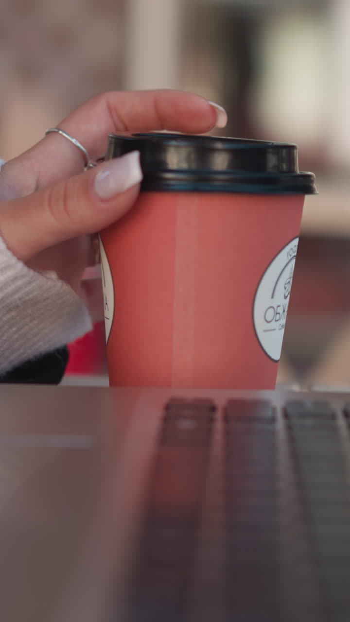 노트북 근처의 커피 컵을 부드럽게 돌리는 여자의 손의 클로즈업, 반지와 함께 그녀의 매니큐어 손가락에 초점을 맞추고, 의자와 함께 흐릿한 배경과 편안한 작업 환경