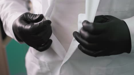 a close-up view of a man wearing black gloves as he carefully un-buttons his lab coat, with a green painted wall behind