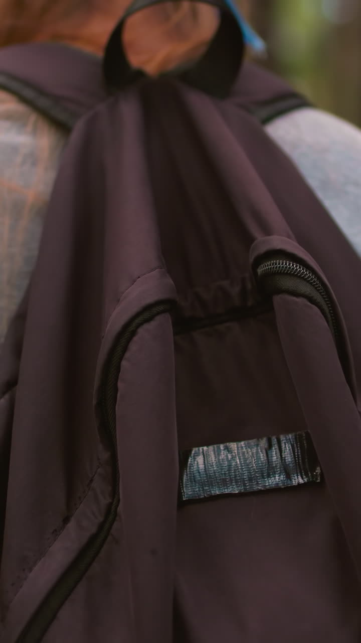 primer plano de una mujer caminando por un sendero forestal con árboles altos en el fondo, balanceando una bolsa negra sobre su hombro, su cabello largo cae en cascada por la espalda