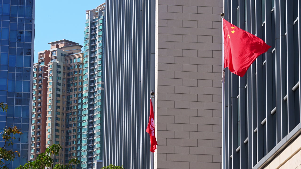 Chinese and Hong Kong flags fluttering on a sunny day in a dense commercial district. The shot illustrates the unique political landscape and urban architecture of the metropolis.
