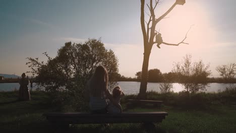 Young-beautiful-mother-and-daughter-are-sitting-on-a-bench-near-a-lake-and-admiring-the-sunset