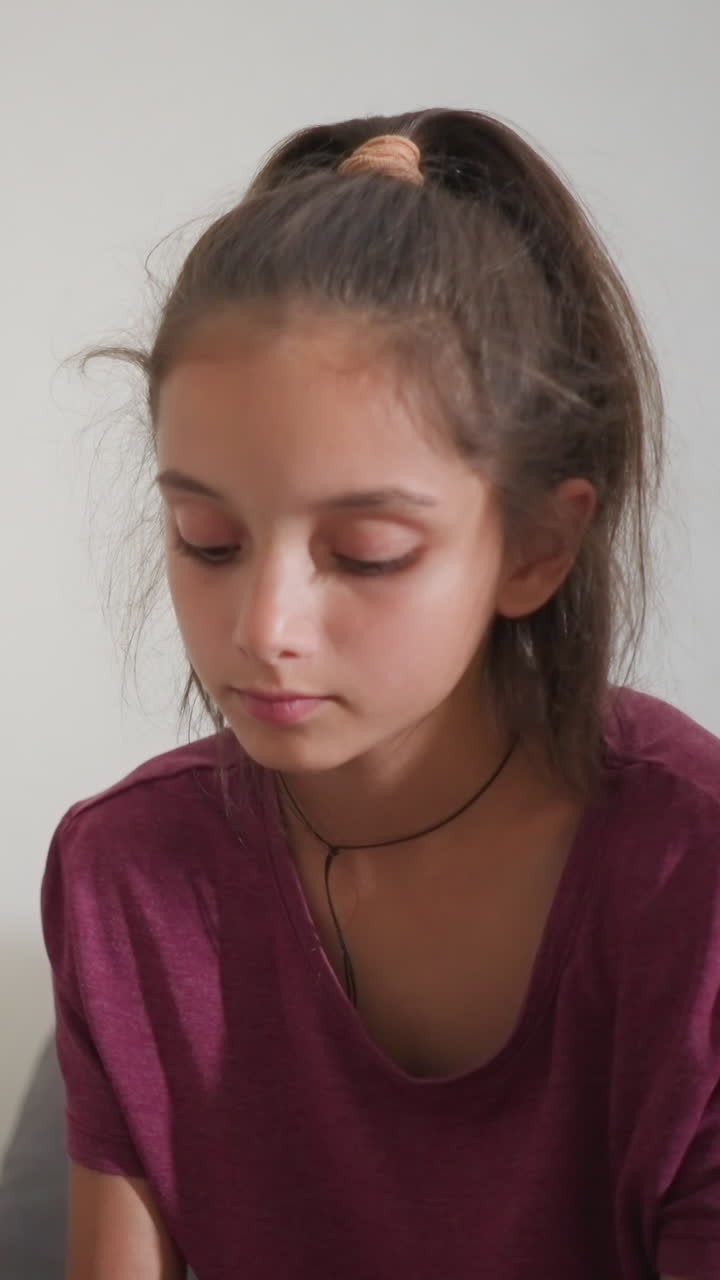 Ragazza con camicia bordeaux seduta in interni che guarda in basso con espressione concentrata mentre una luce soffusa filtra attraverso una tenda trasparente in un ambiente domestico tranquillo con una vista parziale di qualcuno seduto accanto a lei su un comodo divano