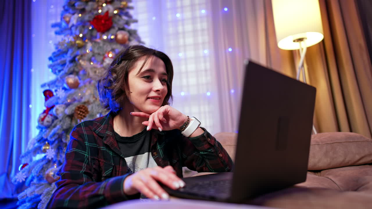 Young woman on laptop, vacation. A young woman sits on a couch, focused on her laptop, with a festive Christmas tree glowing nearby