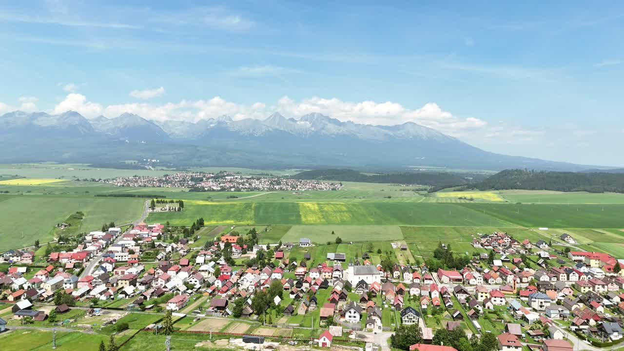 Aerial drone shot slowly flying backward and rotating above green fields and a small church, revealing the vast countryside with the majestic High Tatras mountains in the distance