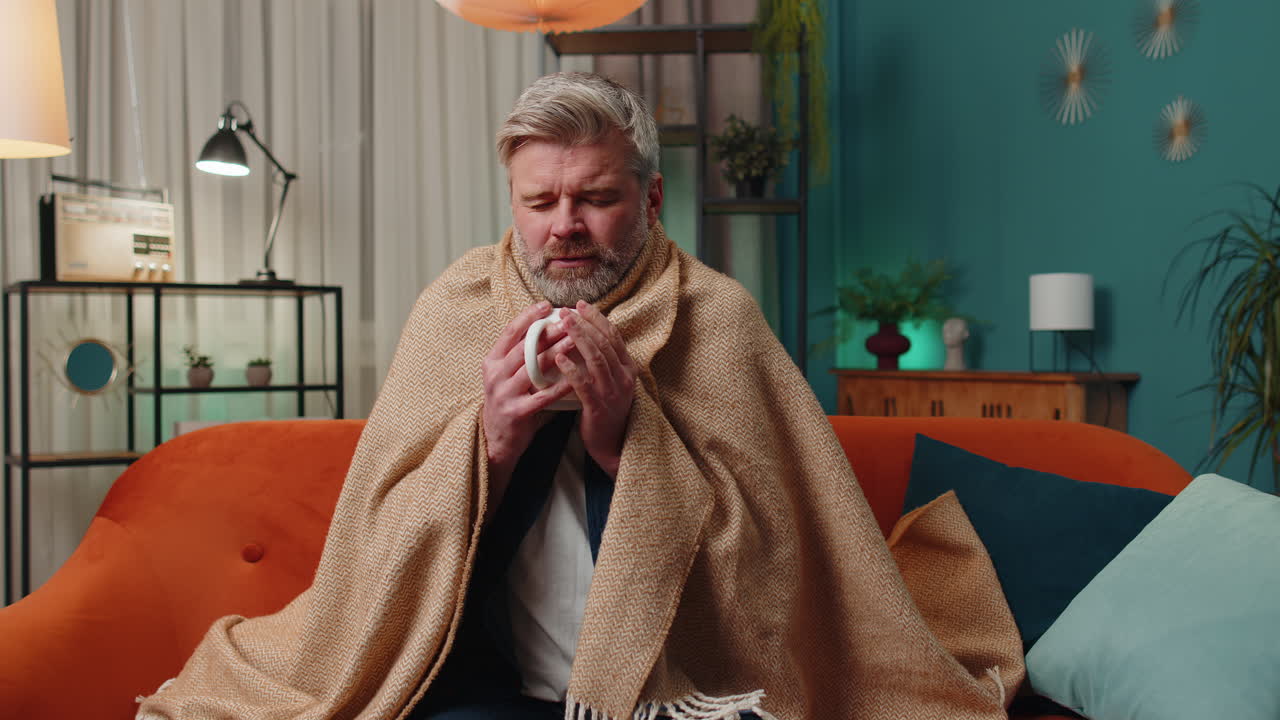 Sick unhealthy man wrapped in plaid sit alone shivering from cold on sofa drinking hot tea at home