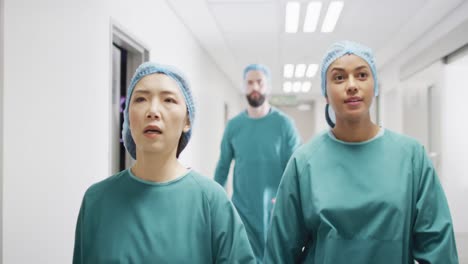 Video-of-four-diverse-surgeons-in-surgical-caps-and-gowns-walking-in-hospital-corridor-talking