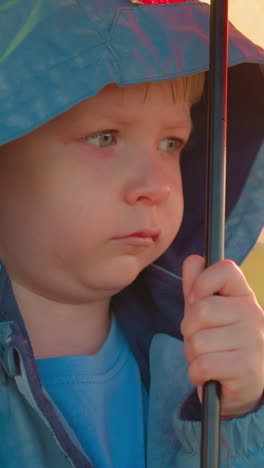 boy stands with parasol in somber mood. child stands alone in green field on rainy day with contemplation punctuated by patter of raindrops