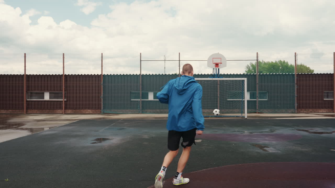 Uomo che si allena a calcio su un campo urbano inzuppato di pioggia con giacca blu, asfalto bagnato e pozzanghere, riscaldamento solitario, poi palleggio e tiro potente verso la porta, cielo nuvoloso e recinzione metallica, grintoso