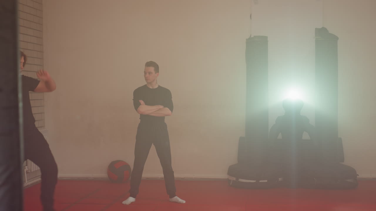 Karate practitioner performing acrobatic cartwheel during martial arts training in gym while partner observes with arms crossed, red mats on floor, punching bags in background, bright spotlight