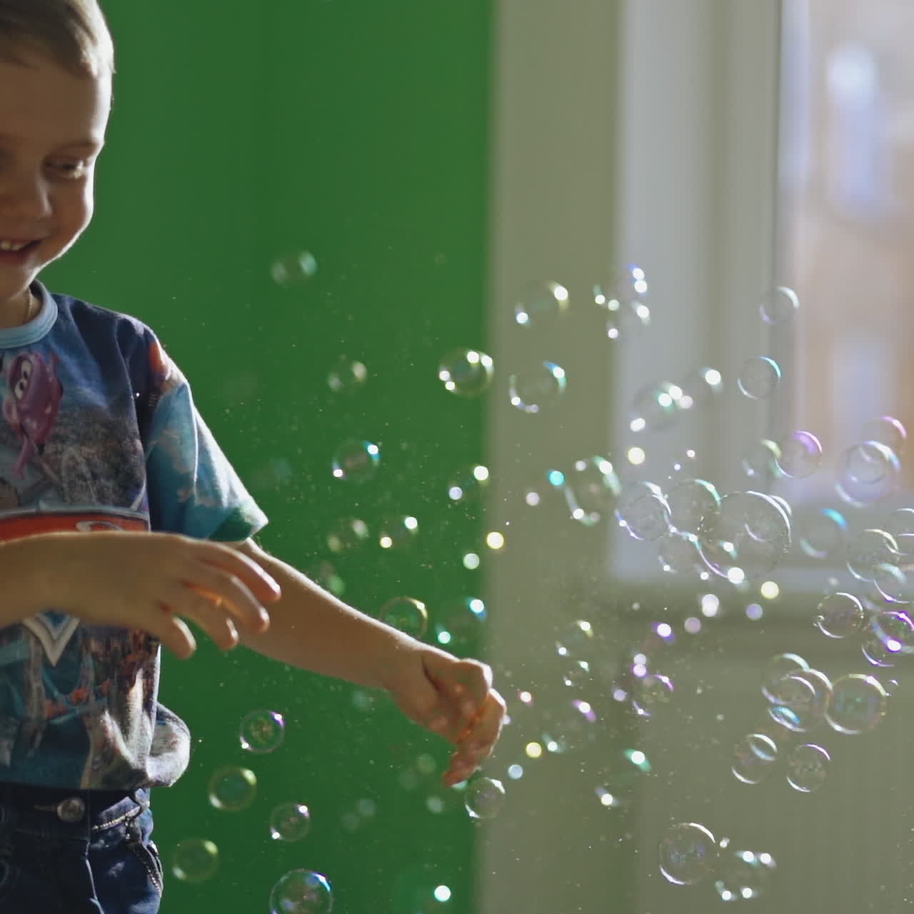 Joyful boy plays with soap bubbles in his room. Little child is popping colorful bubbles with his fingers and smiling at home.