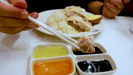 a hand uses chopsticks to dip hainanese chicken into dark soy sauce, with rice and three sauces on a white table under bright lighting