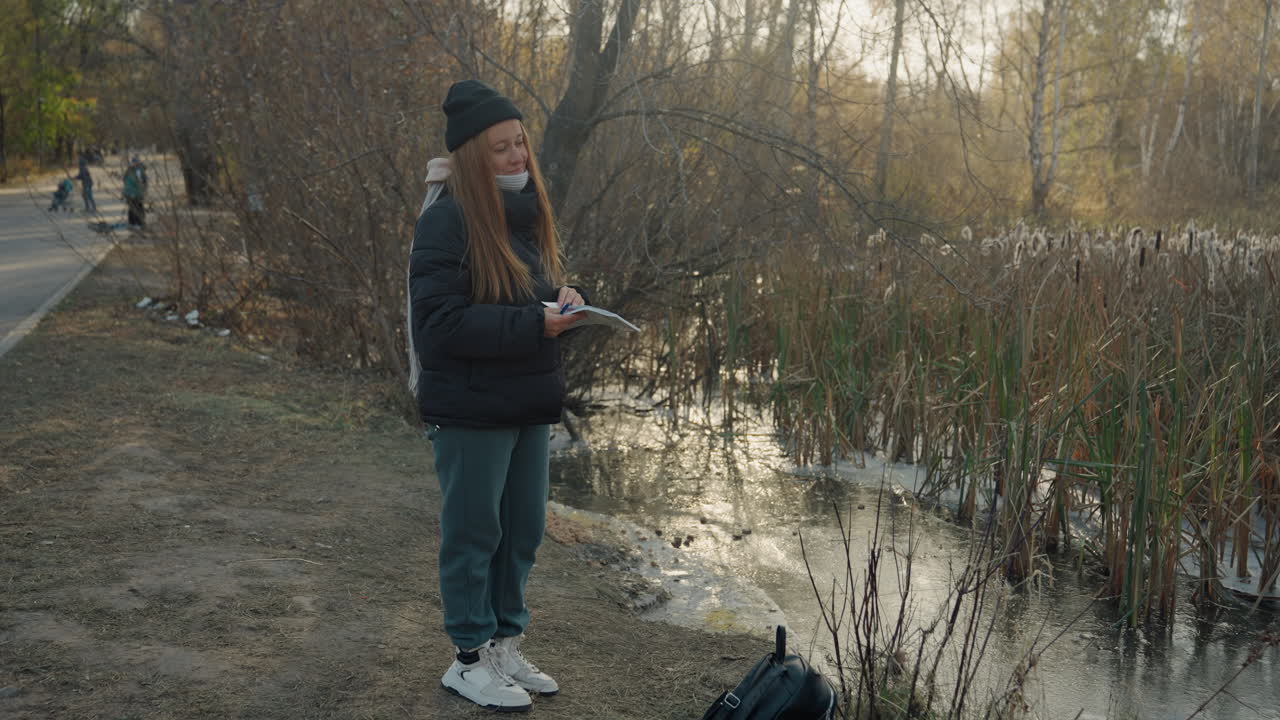 Woman standing by marsh holding controller, late autumn park pathway, puffy jacket and beanie, backpack on ground, reflective pond and reeds, distant pedestrians, quiet tense mood suggesting possible