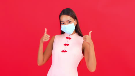 a young woman showing her face mask and gives thumbs up hands gesture, covid-19 virus pandemic concept, red background, static shot