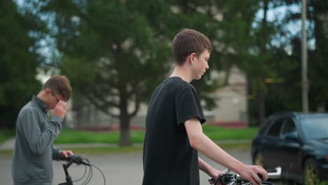 two brothers walking side by side with their bicycles, the one in the grey top adjusts his glasses while the other in a black top walks ahead, the background features trees and a partial view of cars