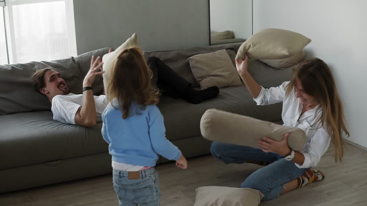 Little cute girl having funny pillow fight with dad on couch in living room and mom is sitting on the floor playing with girl