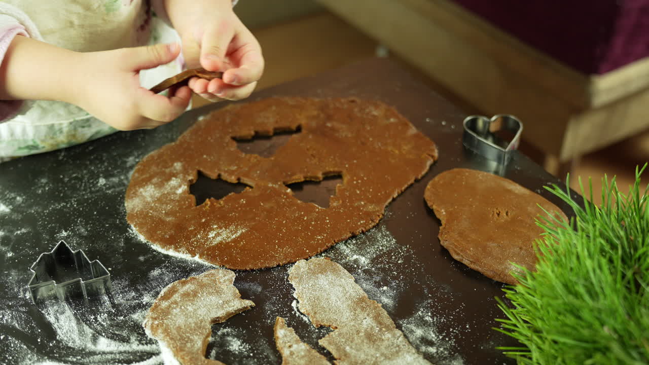 Impasto di pan di zenzero steso viene modellato con formine natalizie da un bambino su un tavolo scuro, con farina sparsa nelle vicinanze e una piccola pianta verde in vista, in un ambiente interno festivo, in tempo reale, inquadratura statica