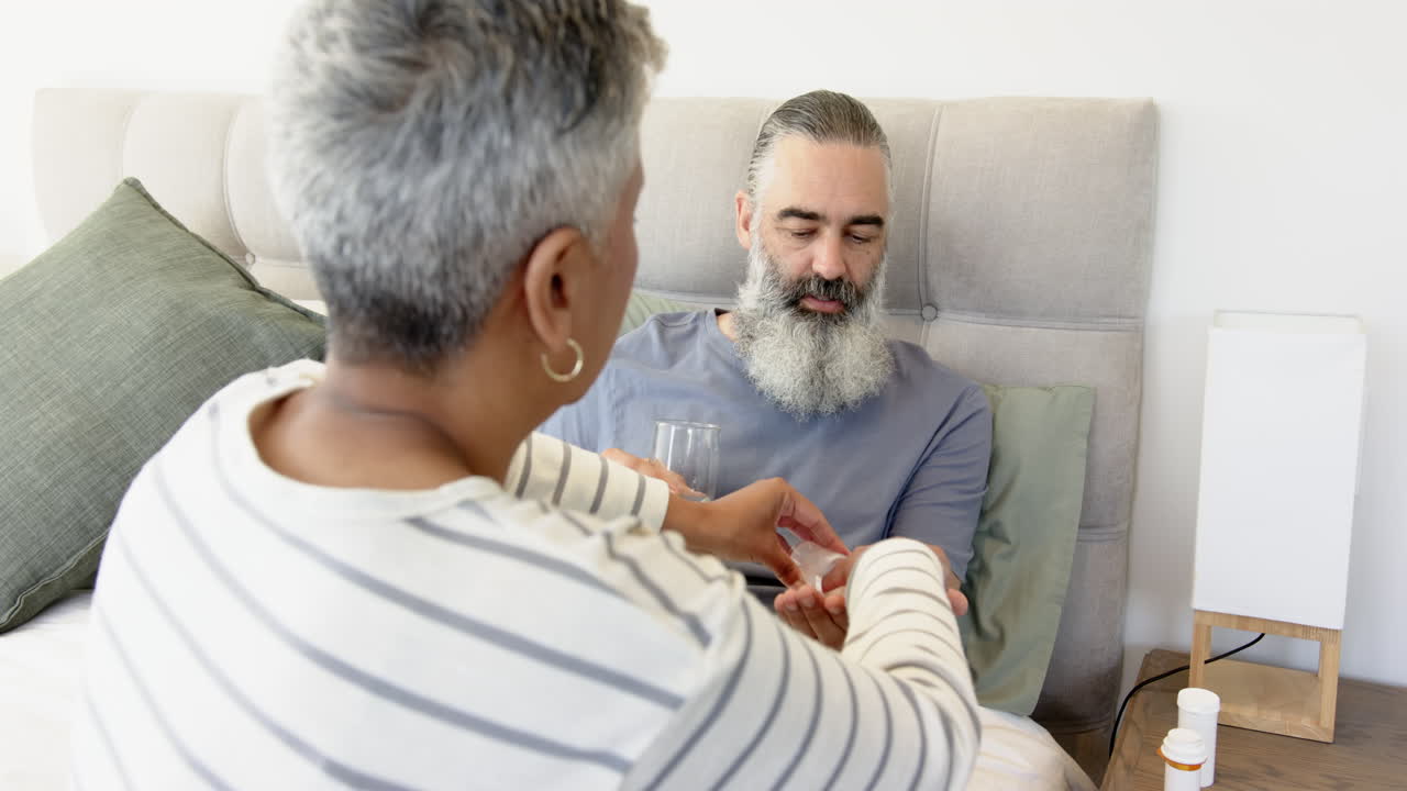Handing medication, woman caring for senior man in bed with glass of water