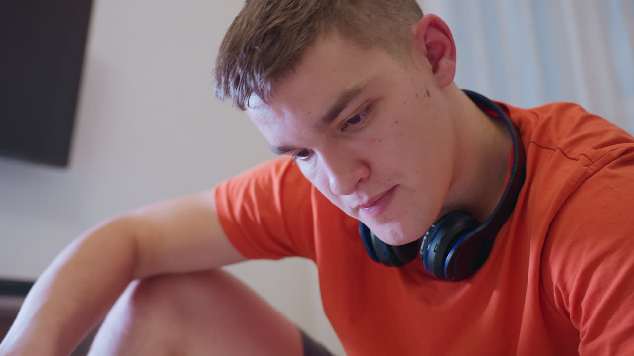 Man in orange shirt seated indoors with black headphones around neck holding screwdriver near chin while appearing thoughtful and reflective in quiet focused moment of concentration and contemplation