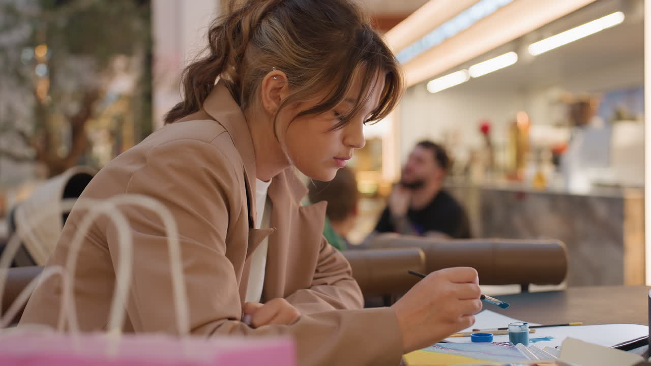 Diseñador con vista lateral dibujando en una cafetería, cuaderno y materiales de pintura en la barra, iluminación cálida, murmullo de fondo y clientes borrosos, planificación enfocada y flujo de trabajo creativo en un entorno público