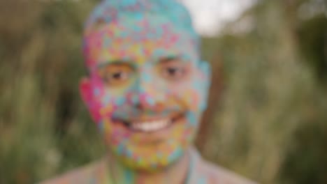 zoom-out portrait of joyful guy standing outdoors at holi festival smiling looking at camera