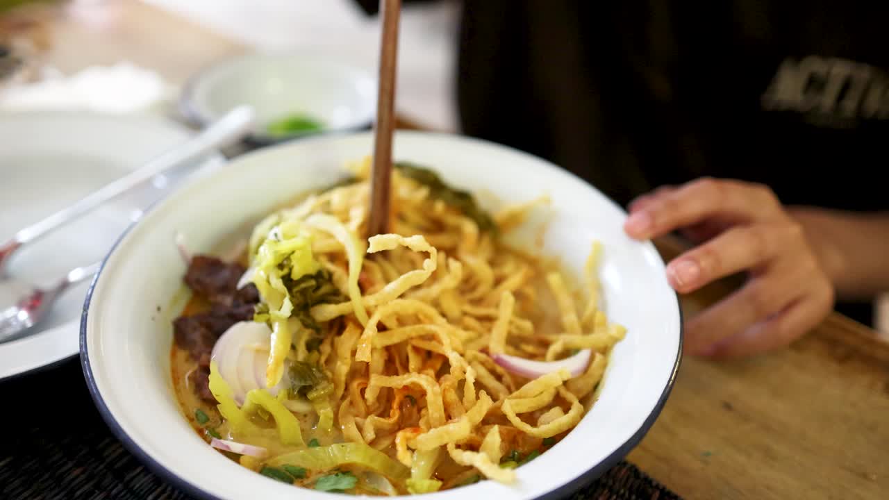 A person uses chopsticks to mix crispy and soft noodles, beef, and vegetables in a bowl of Northern Thai curry, under natural indoor lighting