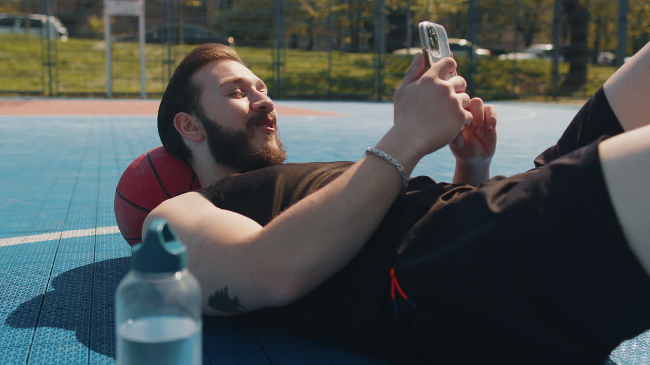 Tired man in sportswear after playing basketball game lying down on ball relaxing using smartphone