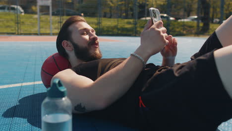 tired man in sportswear after playing basketball game lying down on ball relaxing using smartphone