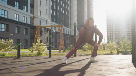 zoom in on resilient woman in hoodie and vest performing side lunge exercise with hands on legs on tiled plaza in urban courtyard framed by autumn foliage and flowers under golden sunlight