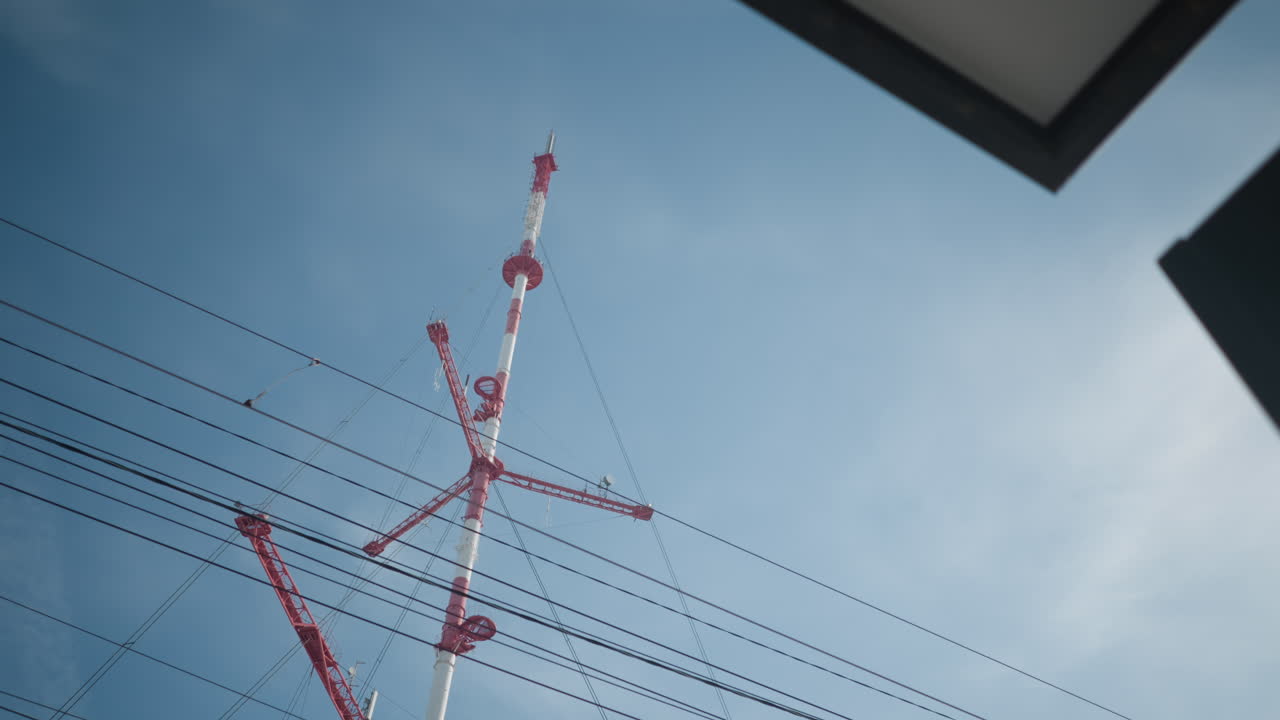upward view past train stop shows red white tower and utility mast with overhead catenary intersecting, blue winter day, urban transit infrastructure humming as power cables web above platform edge