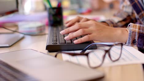de manos femeninas biraciales creativas usando el teclado de la computadora en una oficina casual, cámara lenta
