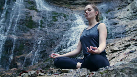 Chica-Practicando-Yoga-Mientras-La-Cascada-Burbujea-En-El-Fondo,-Tiro-De-ángulo-Bajo