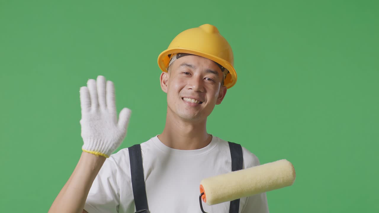 Close Up Of Asian Man Painter Wearing Safety Helmet Smiling, Waving Hand, And Saying Bye While Standing In The Green Screen Background Studio
