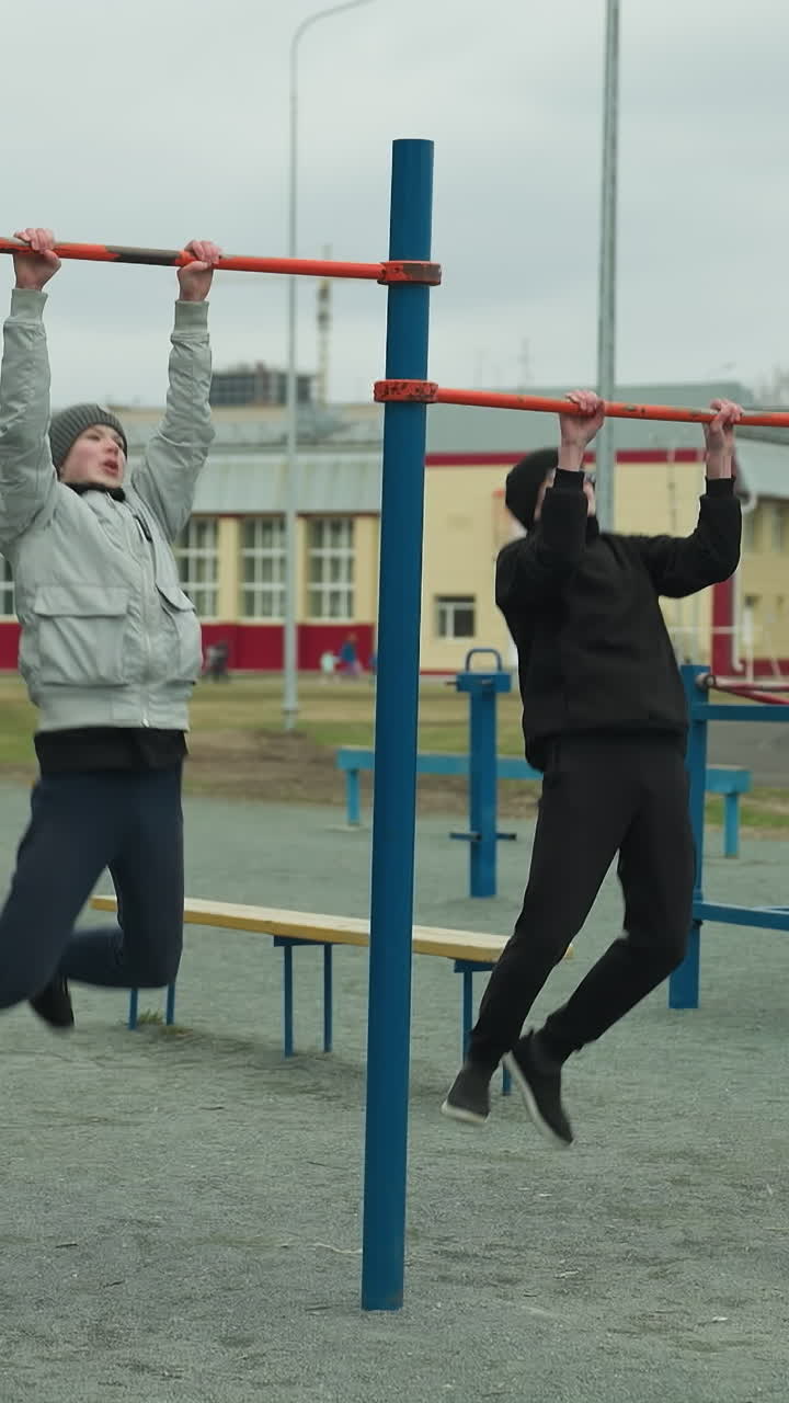 Two boys are working out on pull-up bars in an outdoor gym area, one boy is actively doing a pull-up, while the other joins him, with a man moving around in the background