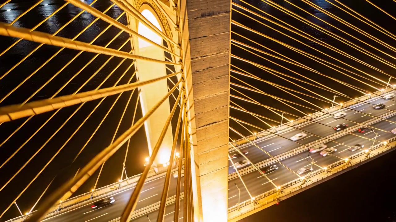 Camera panning central concrete pylon and radiating cables over bridge at night, showing traffic