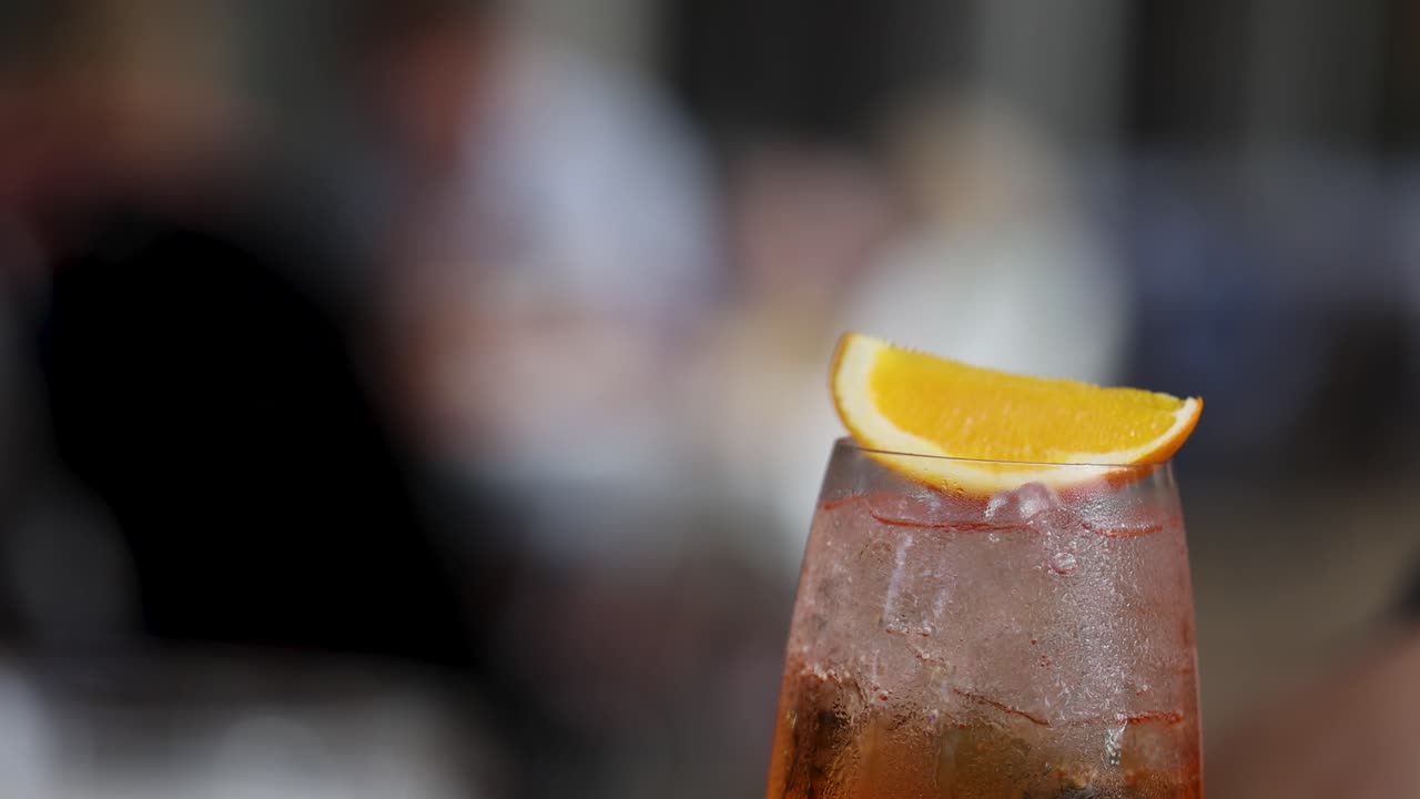 A close-up of an Aperol spritz garnished with a lemon slice, set against a blurred background in warm lighting