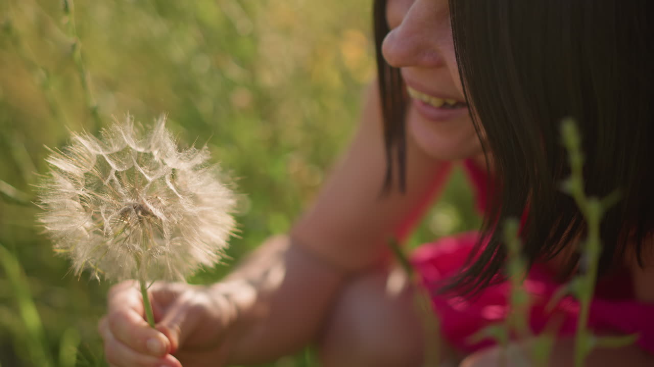 Primer plano de una mujer caucásica en un campo sosteniendo un diente de león mientras sopla las semillas cerca de los labios y la mano con delicada pelusa, luz dorada cálida y bokeh suave, ambiente íntimo y esperanzador de una poetisa o una persona que pide deseos en verano.