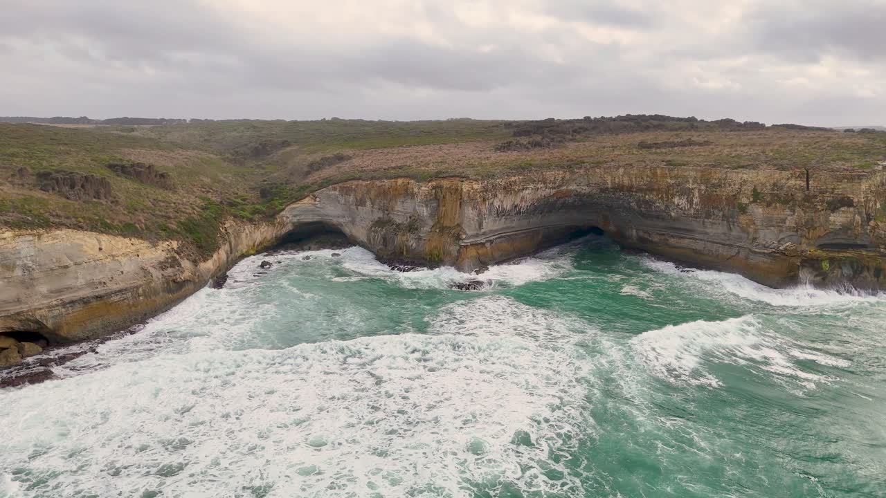 Drone footage captures dramatic cliffs and crashing waves at Port Campbell, Australia. Overcast skies enhance the rugged, natural beauty