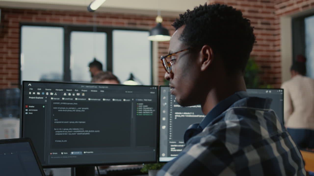 Focused programer fixing glasses while typing on laptop sitting at desk with multiple screens