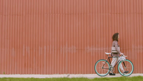 young woman in formal clothes walking with bicycle in front of a prefab metal building