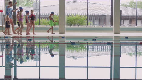 Entrenador-Masculino-Camina-A-Los-Niños-En-Clase-De-Natación-A-Lo-Largo-Del-Borde-De-La-Piscina-Cubierta