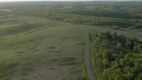 Aerial-shot-of-a-country-road-between-green-meadows-and-a-thick-forest-in-the-mountains-of-Wisconsin,-USA