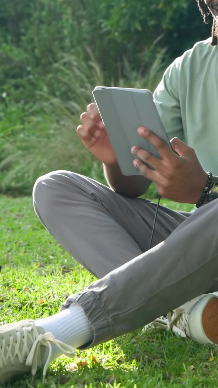 Vertical video: Starting close-up, man in green tee sitting on grass tapping tablet, reacting