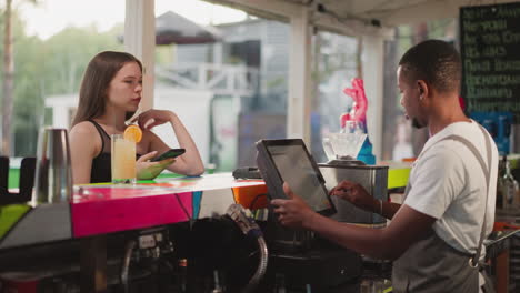 customer waits for bill from barman in cafe. young woman with cocktail and phone opposite black man working on computer in bar. lady buys drink