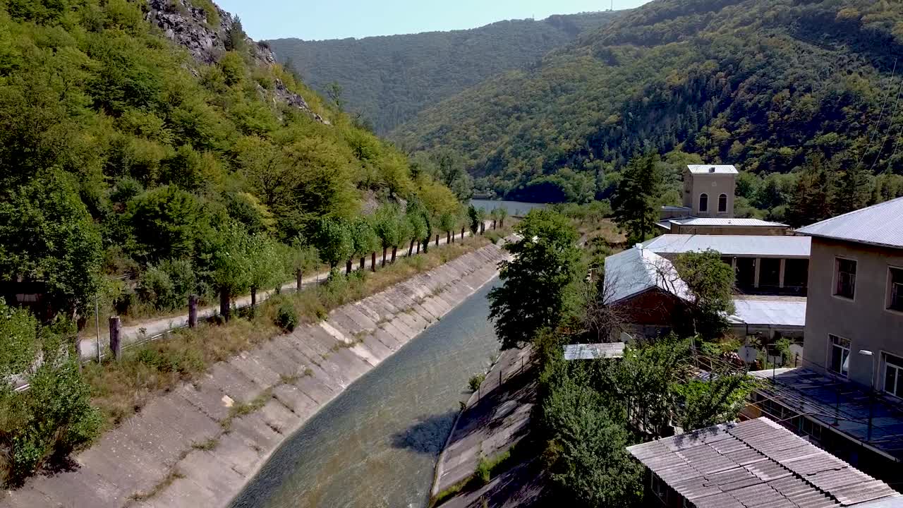 A serene river flows gently between lush green hills and trees, showcasing the peaceful nature of a rural settlement. The sun shines brightly, enhancing the calm atmosphere