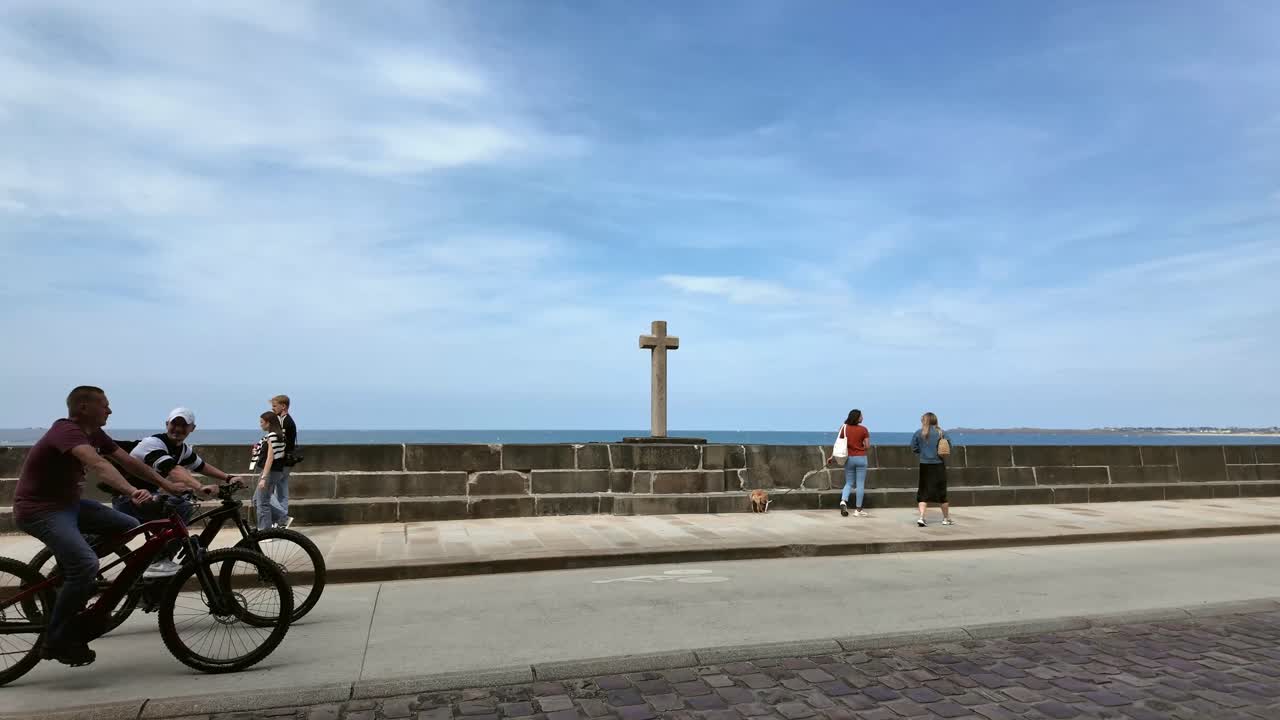 Cyclist passing, two women walking on a coastal promenade with stone cross towards Atlantic, Saint Malo, Brittany, France