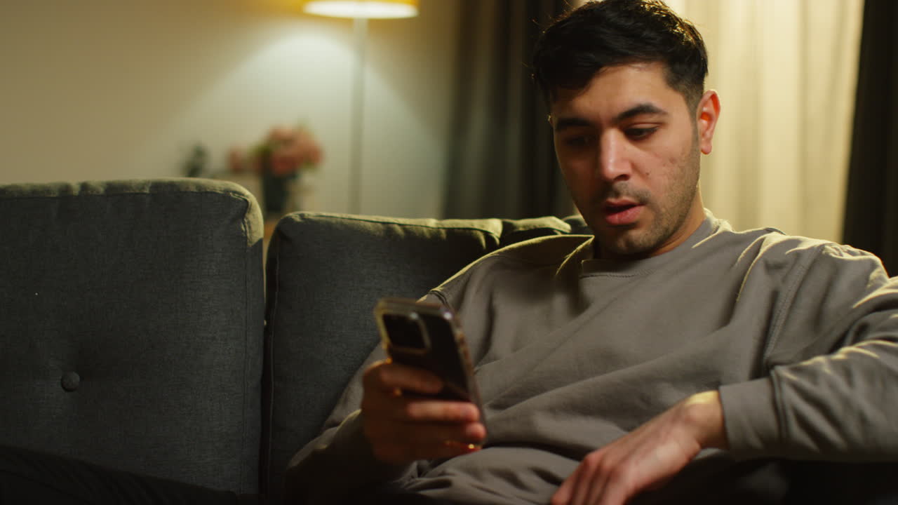 Young Man Spending Evening At Home Sitting On Sofa With Mobile Phone Scrolling Through Internet Or Social Media Streaming Content