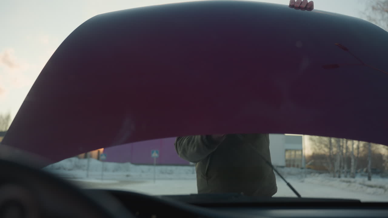 View from inside vehicle shows adult wearing hooded winter jacket lifting car bonnet and positioning support rod to hold it open, with snowy surroundings and distant building visible