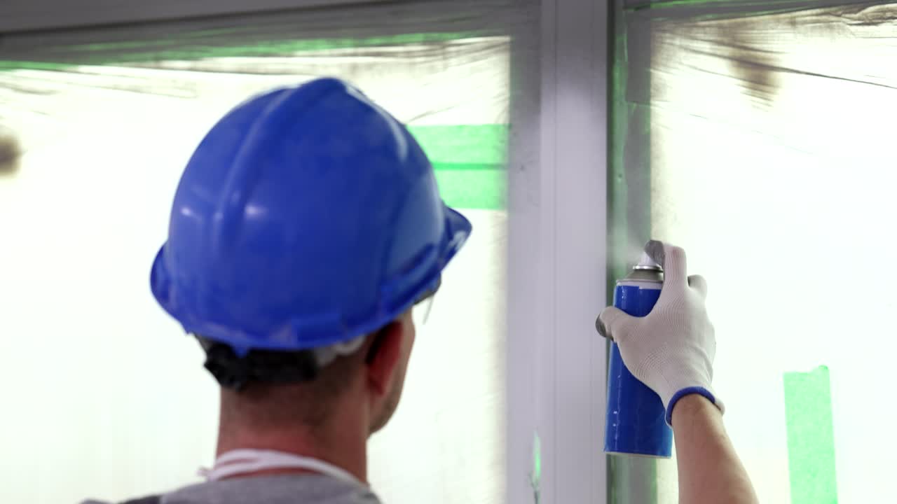 Worker in uniform paints frame with sprayer in apartment
