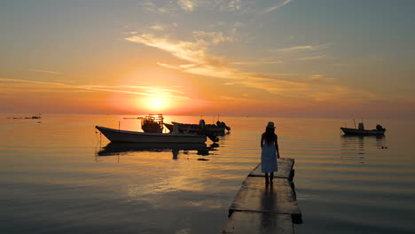 Woman-wearing-white-dress-and-hat-walking-relaxing-on-concrete-bridge-pier-on-the-sea,-with-colorful-sunrise-reflection-sky-and-silhouette-boats-floating-on-water-background,-Bahrain